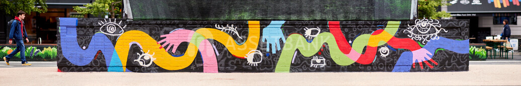 Long outdoor mural featuring bright, flowing shapes and stylised hands in multiple colours on a dark patterned wall.